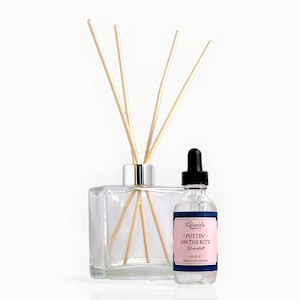May include: A clear glass diffuser with wooden reeds and a small glass bottle with a black dropper. The bottle has a pink label with the text "-PUTTIN' ON THE RITZ Waterfall". The diffuser and bottle are on a white background.
