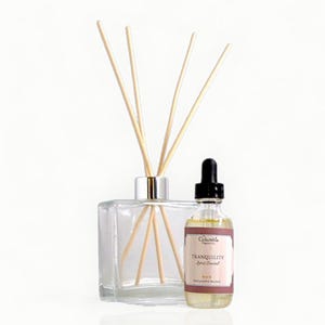 May include: A clear glass diffuser with wooden reeds and a small bottle of essential oil. The oil bottle has a black dropper and a label that reads "Tranquility Label Sandal". The diffuser is square and has a silver collar.