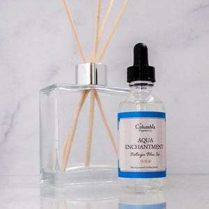 May include: A clear glass bottle with a silver cap and wooden diffuser sticks. The bottle contains a clear liquid and has a blue label that reads "Columbia Fragrance Co. Aqua Enchantment Bellagio Blue Ice Hand poured in Maryland."