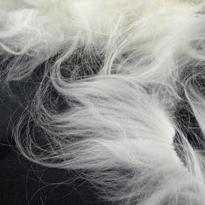 May include: Close-up of white, fluffy fibers, possibly wool or a similar material. The fibers are densely packed and have a soft, textured appearance. The image is well-lit, highlighting the delicate strands and creating a sense of depth.