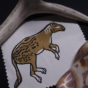 May include: A white fabric square featuring a gold and black block print of a prehistoric animal with a spotted back and striped neck. The fabric has pinked edges and is positioned near a curved antler fragment and a polished brown stone.