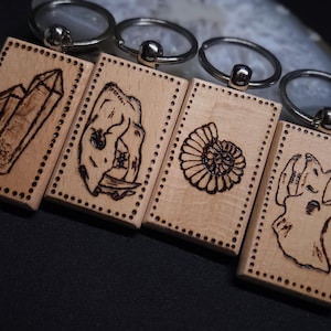 May include: Four rectangular light-colored wooden keychains with silver metal rings. Each keychain features a unique dark wood-burned illustration: a crystal cluster, a skull, an ammonite fossil, and a dinosaur skull, all bordered by dots.