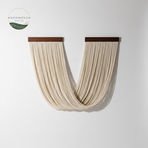 May include: A modern wall hanging featuring cream-colored woven strands suspended from two dark wood planks. The strands curve downward, creating a flowing, artistic design. The piece is mounted on a white wall, with the Handwoven Nature logo in the upper left corner.
