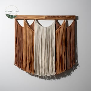 Op de afbeelding: Handgemaakte macramé wandhanger met een houten stok en drie franjes in bruin- en crèmetinten. De wanddecoratie heeft een halfronde vorm en voegt een vleugje bohemien stijl toe aan elke kamer.