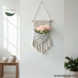 Puede incluir: Un colgante de pared de macramé color crema con una clavija de madera y un cordón colgante triangular. El bolsillo tejido contiene flores rosas y vegetación artificial. La decoración de la pared presenta flecos en la parte inferior. El texto "HANDWOVEN NATURE".