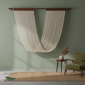 Op de afbeelding: Crèmekleurige, geweven gordijnpalen die aan een donkerbruine stang hangen, waardoor een gedrapeerd effect ontstaat tegen een gedempte groene muur. Een jute vloerkleed en een groene fauteuil maken het interieur compleet. Het logo "HANDWOVEN NATURE" is zichtbaar.