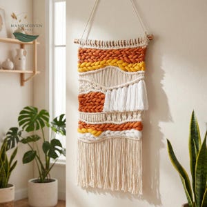 Op de afbeelding: Handgeweven wandkleed met een natuurlijk kleurenpalet. Het textielkunstwerk heeft een mix van texturen, waaronder geweven delen, geknoopte details en franjes. Kleuren zijn tinten oranje, geel, crème en wit. Het is opgehangen aan een houten deuvel.