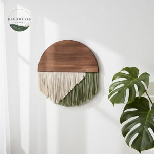 May include: A round wall hanging with a dark wood top and a textured, fringed bottom. The bottom is divided into two sections: off-white and olive green. The wall hanging is mounted on a white wall, with a green plant in the background.
