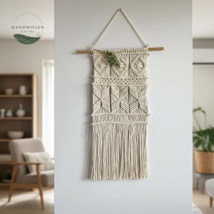 May include: Handwoven macrame wall hanging in cream color, featuring geometric patterns and fringe. The wall decor is suspended from a wooden dowel with a cream-colored cord. The logo "HANDWOVEN NATURE" is visible in the upper left corner.