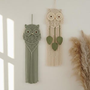 May include: Two macrame owl wall hangings. One is sage green, the other is cream. Both have wooden bead eyes and leaf accents. The green owl has long fringe. The cream owl has three green leaf accents hanging from the bottom.