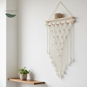 May include: A cream macrame wall hanging with a wooden shelf and a small woven basket. The macrame design features a geometric pattern. The wall hanging is suspended from a wooden dowel. A wooden shelf and a small potted plant are also visible.