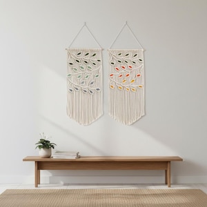 以下が含まれることがあります： 葉のデザインが施された、2つのマクラメの壁掛け。各作品は、緑、青、黄、オレンジの色合いの葉のシリーズを備えたクリーム色の背景が特徴です。壁掛けは木のダボから吊り下げられています。