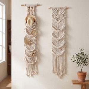 May include: Two macrame wall organizers in a natural cream color. One holds a straw hat, dried palm leaves, and twine. The other features a geometric design. Both are suspended from wooden dowels, adding a touch of bohemian decor.