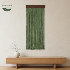 Pode incluir: Uma tapeçaria de parede tecida verde com uma barra de madeira marrom escura. A tapeçaria de parede é feita de muitos fios verticais. A imagem também mostra um banco de madeira e um vaso branco com uma planta. O logotipo "HANDWOVEN NATURE" está no canto superior esquerdo.