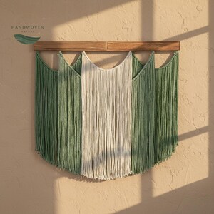 Puede incluir: Un tapiz de macramé con una barra de madera en la parte superior y flecos verdes y blanquecinos. Los flecos están dispuestos simétricamente, con la sección blanquecina en el centro. Se puede ver la etiqueta "HANDWOVEN NATURE".