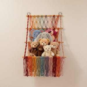 May include: A macrame wall shelf with a rainbow design, holding a collection of plush toys. The toys include teddy bears, an elephant, a bunny, and a pink bear. The shelf is made of wood and colorful yarn.