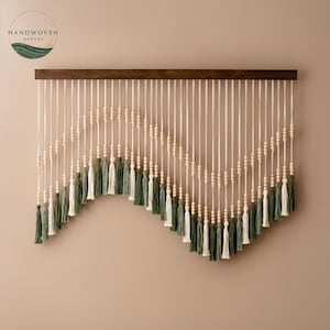 Puede incluir: Un colgante de pared artesanal con cuerdas blancas, cuentas de madera y borlas verdes y blancas. El diseño está montado sobre una barra de madera marrón oscuro, creando una pieza decorativa inspirada en la naturaleza para la decoración del hogar.