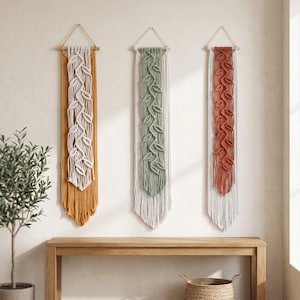 May include: Three macrame wall hangings with leaf patterns in mustard yellow, sage green, and rust orange. Each piece has a wooden dowel and a triangular top for hanging. The wall hangings are displayed on a neutral wall.
