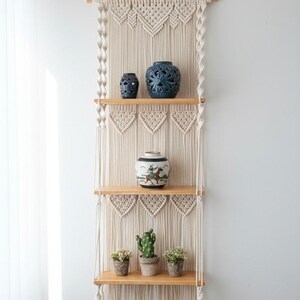 May include: A macrame wall shelf with three wooden shelves. The shelf is decorated with a variety of items, including blue ceramic vases, a decorative jar, and small potted plants. The macrame design is cream-colored.