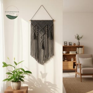 May include: A gray macrame wall hanging with a geometric design and tassels. The wall decor is suspended from a wooden dowel and features the text "HANDWOVEN NATURE". The macrame is hanging on a white wall.