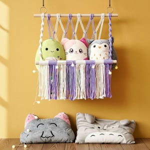 May include: A macrame wall hanging with three plush animal pillows: a green avocado, a pink cat, and a gray owl. Below, two gray character pillows, one cat-shaped and one resembling a cartoon character, rest on the surface. The wall is yellow.