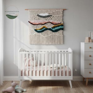 May include: A white crib with a pink sheet and a woven wall hanging with colorful, textured layers. The wall hanging features cream, pink, gray, teal, and gold tones. The image also includes a white dresser and two stuffed animals.