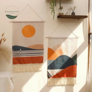 May include: Two rectangular wall hangings with landscape designs. One features an orange sun over blue and tan hills, a sailboat, and a fringe. The other has an orange sun over mountains with a fringe. The brand logo "HANDWOVEN NATURE" is in the upper left.