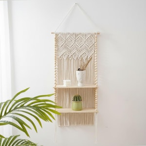 Puede incluir: Una estantería de pared de macramé con dos estantes de madera. La sección superior presenta un patrón de diamantes tejido y flecos colgantes. Los estantes contienen un pequeño cactus en una maceta verde, un jarrón blanco con forma de cara y velas.
