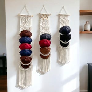May include: Three macrame hat organizers hanging on a white wall. Each organizer has a wooden dowel top and holds several baseball caps in various colors, including brown, navy, and red. The organizers have a fringe detail at the bottom.
