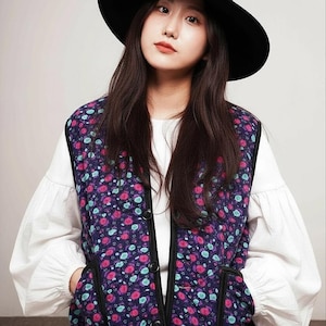 Pode incluir: Uma mulher usando um chapéu preto e um colete com estampa floral sobre uma blusa branca. O colete apresenta um fundo roxo escuro com padrões florais rosa e verde-azulado. A roupa é completada com calças brancas.