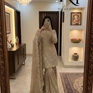 Puede incluir: Un conjunto salwar kameez beige con intrincados adornos de lentejuelas. El atuendo incluye una túnica de manga larga, pantalones de pernera ancha y una dupatta a juego. El texto "RIVARA INDIA" es visible.