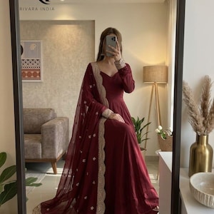 Puede incluir: Un vestido formal de color burdeos con mangas largas y falda fluida. El vestido presenta un escote en V y se combina con una bufanda transparente a juego con ribete dorado. También se muestra el texto "RIVARA INDIA".