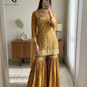 May include: Mustard yellow embroidered Indian salwar kameez set. The outfit includes a long-sleeved tunic, wide-legged pants, and a matching scarf. The set features intricate gold embroidery and decorative trim. The brand logo "RI RIVARA INDIA" is visible in the top left corner.
