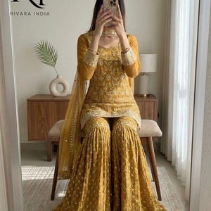 May include: Mustard yellow embroidered salwar kameez set. The set includes a long-sleeved top, wide-legged pants, and a matching scarf. The outfit features intricate gold embroidery and is from Rivara India.