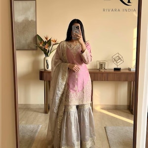 May include: A pink embroidered salwar kameez set with matching dupatta. The outfit features intricate gold embroidery on the neckline, sleeves, and hem. The palazzo pants are a light grey colour. The mirror reflects the text "RIVARA INDIA".