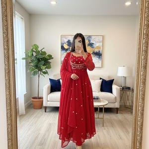 Könnte beinhalten: Ein leuchtend rotes Kleid mit aufwendiger Stickerei am Oberteil und verstreuten goldenen Verzierungen. Das langärmlige Kleid enthält einen passenden Schal. Das Outfit wird mit goldfarbenen Stiefeln kombiniert. Das Kleid ist für besondere Anlässe geeignet.