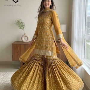 May include: Mustard yellow Indian salwar kameez set. The outfit features a long-sleeved tunic with intricate embroidery and flared pants. A matching scarf completes the ensemble. The outfit is embellished with silver accents and is suitable for festive occasions.
