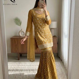 May include: Mustard yellow embroidered salwar kameez set. The outfit includes a long-sleeved tunic, flared pants, and a matching scarf. The ensemble features intricate embroidery and a gold trim. The brand logo "RI RIVARA INDIA" is visible in the top left corner.