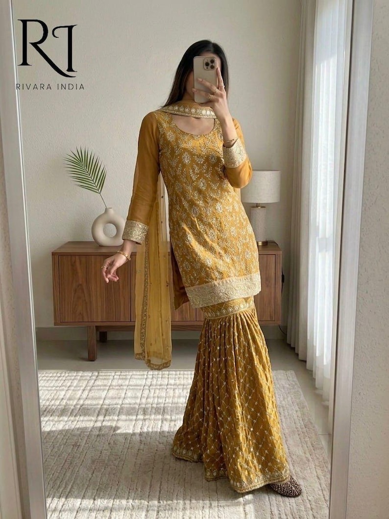 May include: Mustard yellow embroidered salwar kameez set. The outfit includes a long-sleeved tunic, flared pants, and a matching scarf. The ensemble features intricate embroidery and a gold trim. The brand logo "RI RIVARA INDIA" is visible in the top left corner.