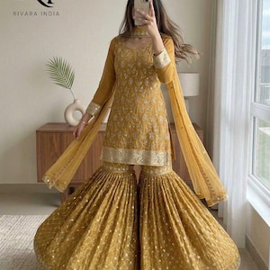 May include: Mustard yellow embroidered Sharara suit with a matching dupatta. The outfit features intricate detailing on the bodice and flared pants. The sleeves and edges of the dupatta are adorned with gold trim. The outfit is from Rivara India.