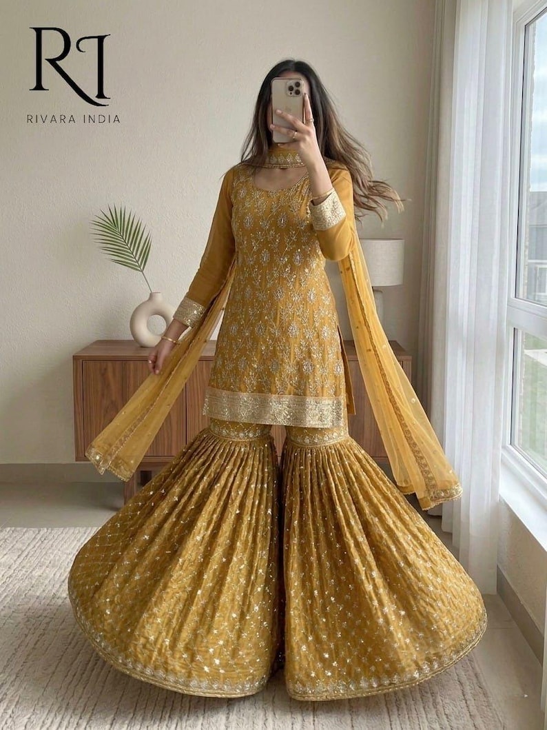 May include: Mustard yellow embroidered Sharara suit with a matching dupatta. The outfit features intricate detailing on the bodice and flared pants. The sleeves and edges of the dupatta are adorned with gold trim. The outfit is from Rivara India.