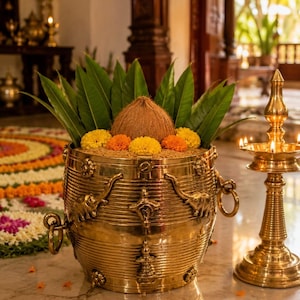 May include: A polished brass vessel featuring embossed elephant motifs and side ring handles, filled with grain, green leaves, a brown coconut, and yellow and orange marigold flowers, positioned next to a lit brass oil lamp.