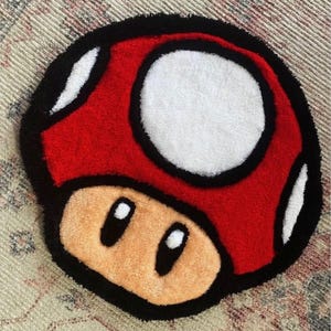 May include: A mushroom-shaped rug in red, white, black, and tan. The rug has a red cap with white spots, a white circle, and a tan base with black eyes. This playful rug is a decorative item.
