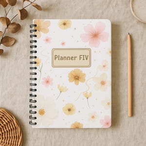 Peut inclure: Un carnet à spirales avec une couverture blanche ornée de fleurs aux tons pêche, roses et jaunes. Le texte "Planner FIV" est affiché dans un cadre rectangulaire. Un crayon en bois est posé à côté du carnet.