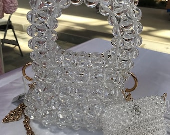 May include: A clear beaded handbag with a matching small purse. The handbag has a circular handle and a gold chain strap. The purse is rectangular and also has a gold chain. Both are made of clear, round beads.