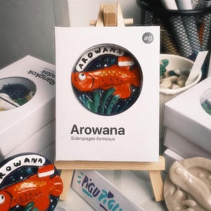 Może przedstawiać: Biała prostokątna karta prezentacyjna z okrągłym wycięciem, w którym znajduje się wyrzeźbiona pomarańczowa figurka ryby Arowana. Na karcie widnieje napis Arowana, Scleropages formosus oraz numer #6. Karta spoczywa na małej drewnianej sztaludze.