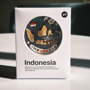 Puede incluir: Una caja rectangular blanca con un recorte circular que muestra un mapa en relieve de Indonesia en oro sobre un fondo azul oscuro. La caja incluye el texto #1, Indonesia, y una descripción del país como un archipiélago tropical.