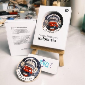 Puede incluir: Un broche redondo de cerámica con forma de máscara de Cirebon, de rostro rojo con tocado y bigote azul oscuro, etiquetado como Indonesia. Se exhibe en un pequeño caballete de madera junto a una caja blanca y una tarjeta descriptiva.