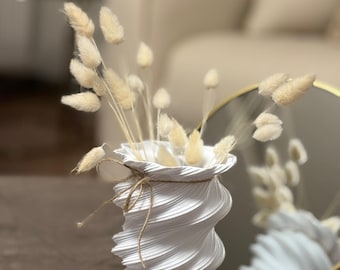 May include: A white, spiral-shaped vase with dried, beige bunny tail grass. The vase has a textured surface and is tied with twine around the rim. A gold-framed mirror reflects the vase and grass. The vase is on a dark brown surface.