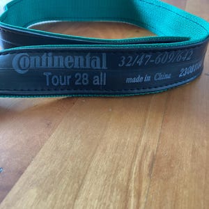 Könnte beinhalten: Ein schwarzer Continental Tour 28 all Fahrradreifen mit grünem Stofffutter. Der Reifen trägt die Aufschrift "Continental" und "Tour 28 all" sowie Größen- und Herstellungsinformationen. Der Reifen wird in China hergestellt.